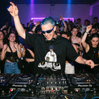 DJ with blue hair and sunglasses wearing Chill Session DJ graphic oversized t-shirt performing live with arms raised