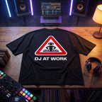 Black oversized t-shirt with DJ At Work warning sign graphic, flat lay on wooden desk with DJ controller and vinyl records
