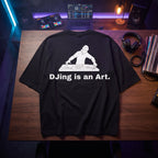Black oversized t-shirt with DJing Is An Art graphic print, flat lay on wooden desk with DJ controller and vinyl records