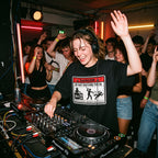 A woman DJing at a nightclub or party, wearing a black t-shirt with a 'Danger Do Not Disturb' graphic. She is smiling and waving, with her hands in the controls of a Pioneer DJ turntable.
