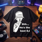 Black oversized t-shirt with Shh He's The Best DJ graphic print, flat lay on wooden desk with vinyl records and DJ controller
