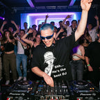 DJ with blue hair and sunglasses wearing Shh He's The Best DJ oversized black t-shirt performing at nightclub