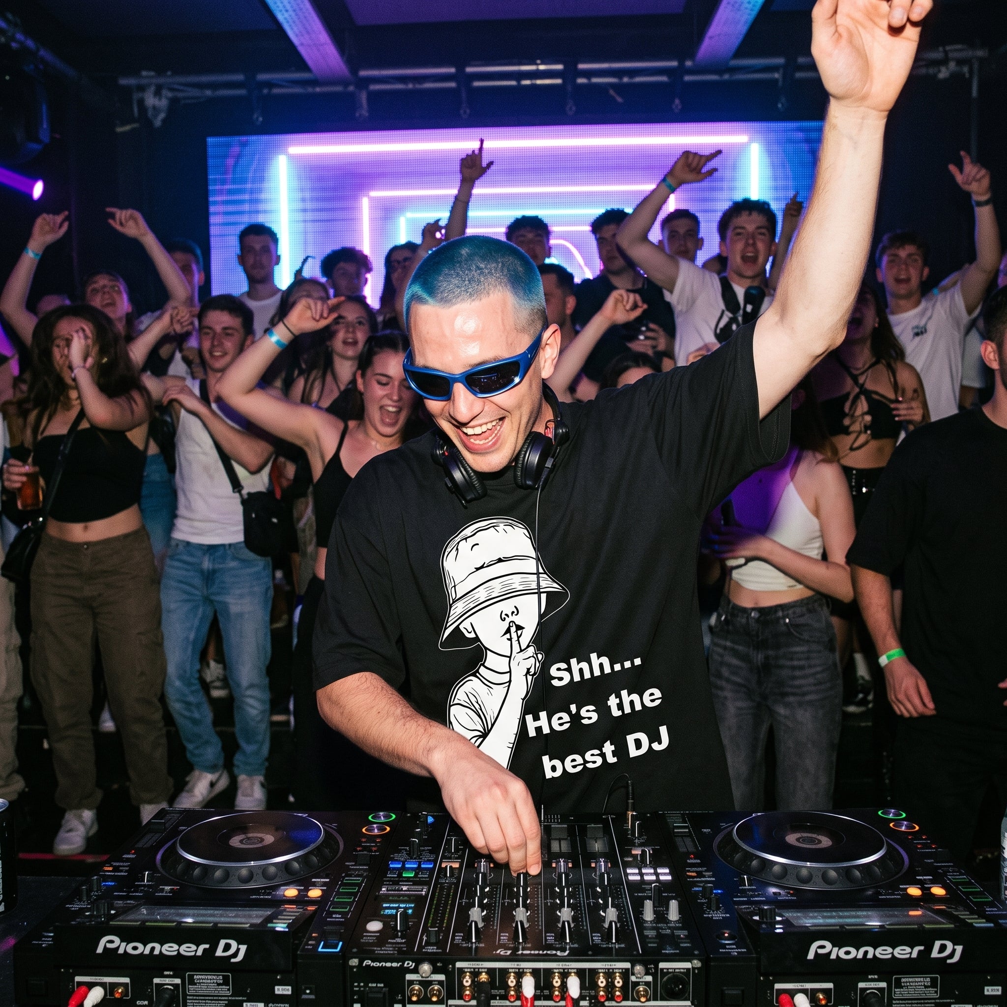 DJ with blue hair and sunglasses wearing Shh He's The Best DJ oversized black t-shirt performing at nightclub