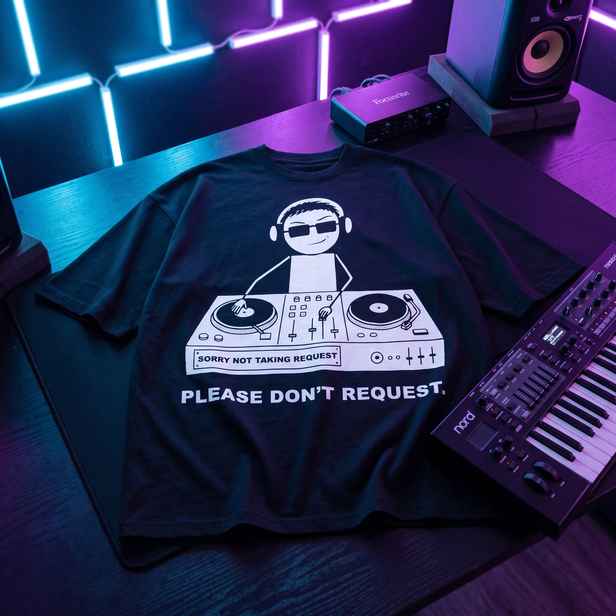 Black oversized DJ t-shirt with Please Don't Request graphic laid flat under neon blue and purple studio lighting