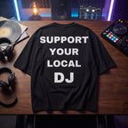 Black oversized t-shirt with Support Your Local DJ print, flat lay on wooden desk with DJ controller and vinyl records