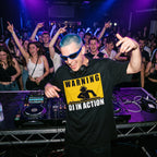 DJ with blue hair and sunglasses wearing Warning DJ In Action oversized black t-shirt performing at nightclub