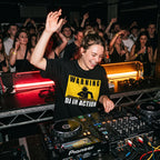 Female DJ wearing Warning DJ In Action oversized t-shirt mixing on decks with energetic crowd under red lights