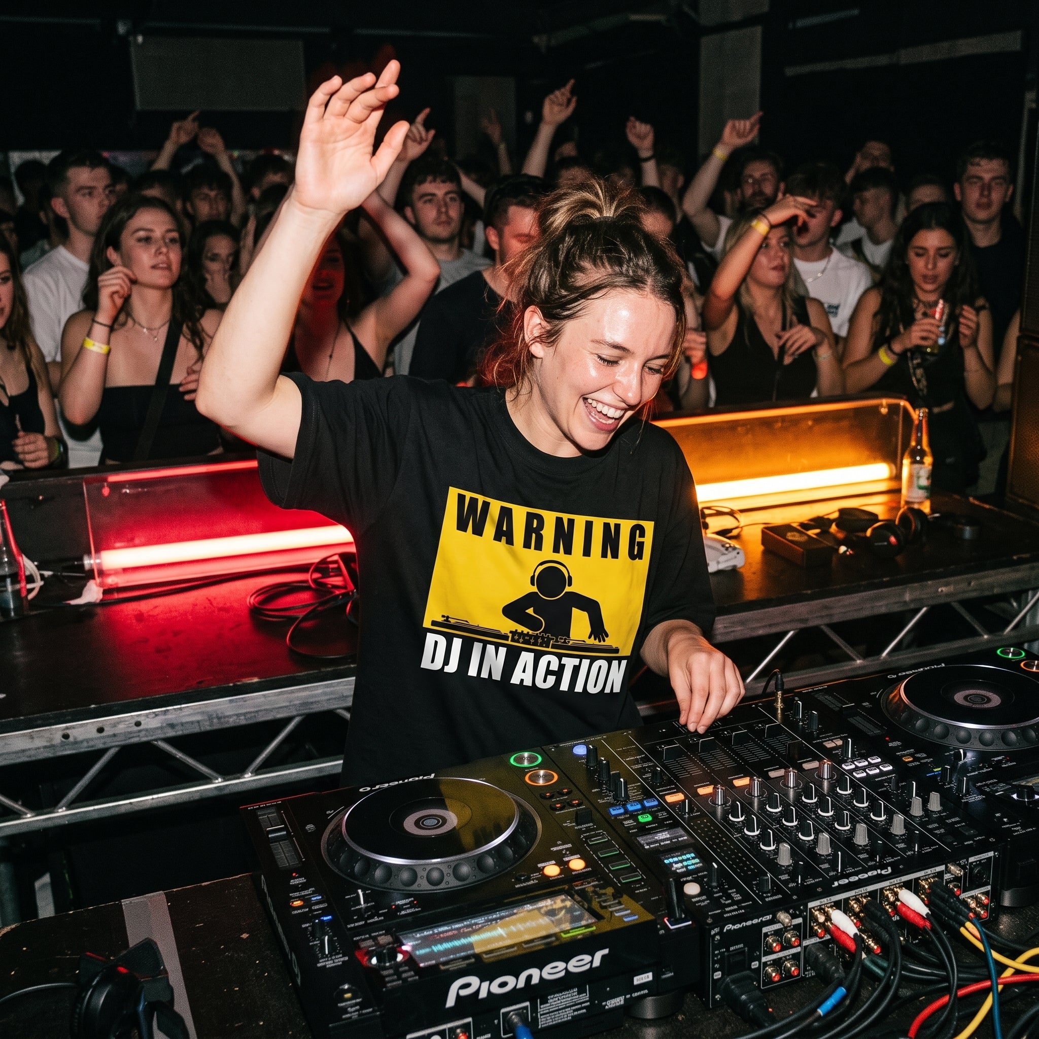 Female DJ wearing Warning DJ In Action oversized t-shirt mixing on decks with energetic crowd under red lights