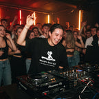 Female DJ wearing World's Best DJ quote oversized t-shirt mixing on decks with energetic crowd under red lights