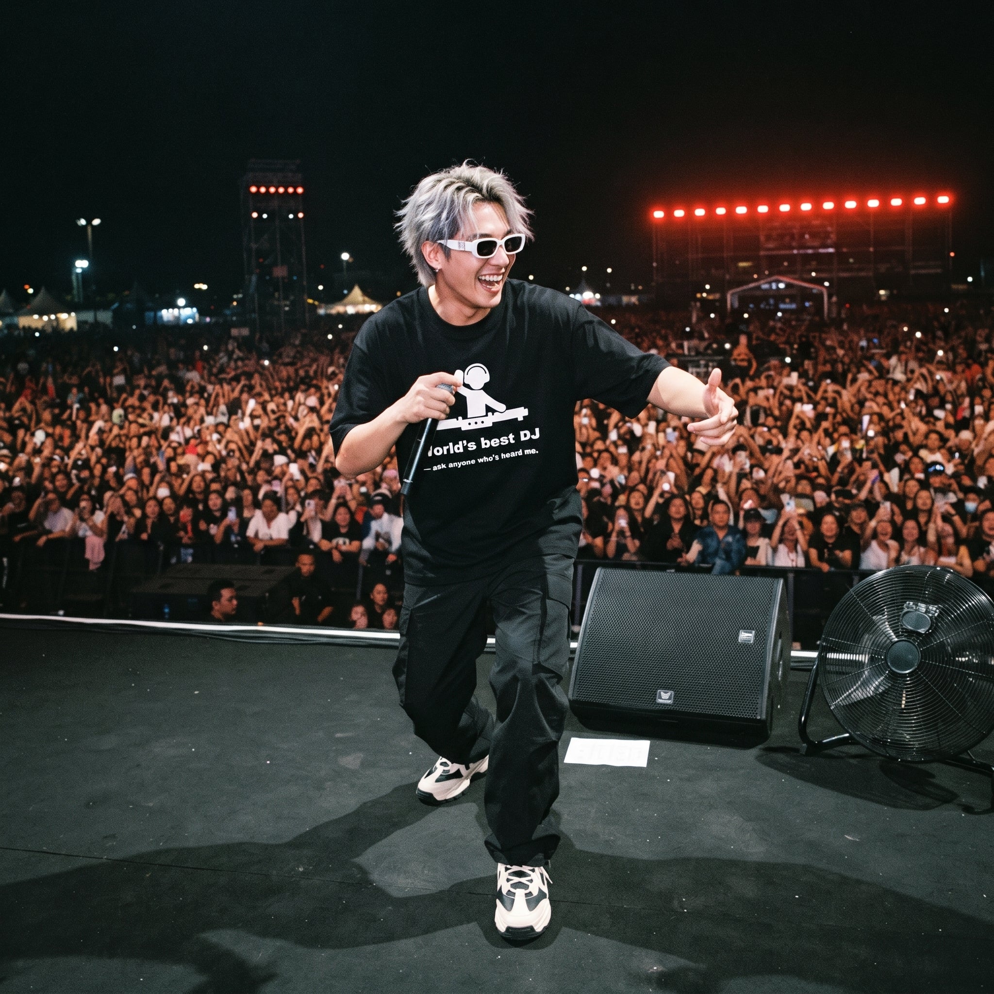 DJ in World's Best DJ quote oversized black t-shirt holding microphone on outdoor festival stage in front of large crowd