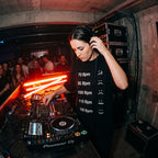 Female DJ in BPM Chart Oversized T-Shirt adjusting headphones at DJ booth club